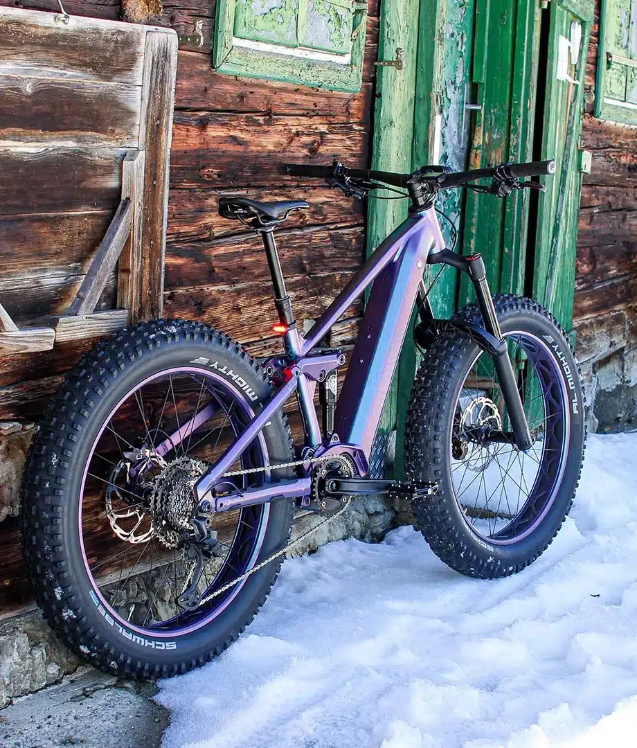 Full-Suspension E-Mountainbike MAXX Huraxdax ELS X2 mit extra breiten Reifen, abgestellt im Schnee vor einer rustikalen Holzhütte mit grüner Tür.