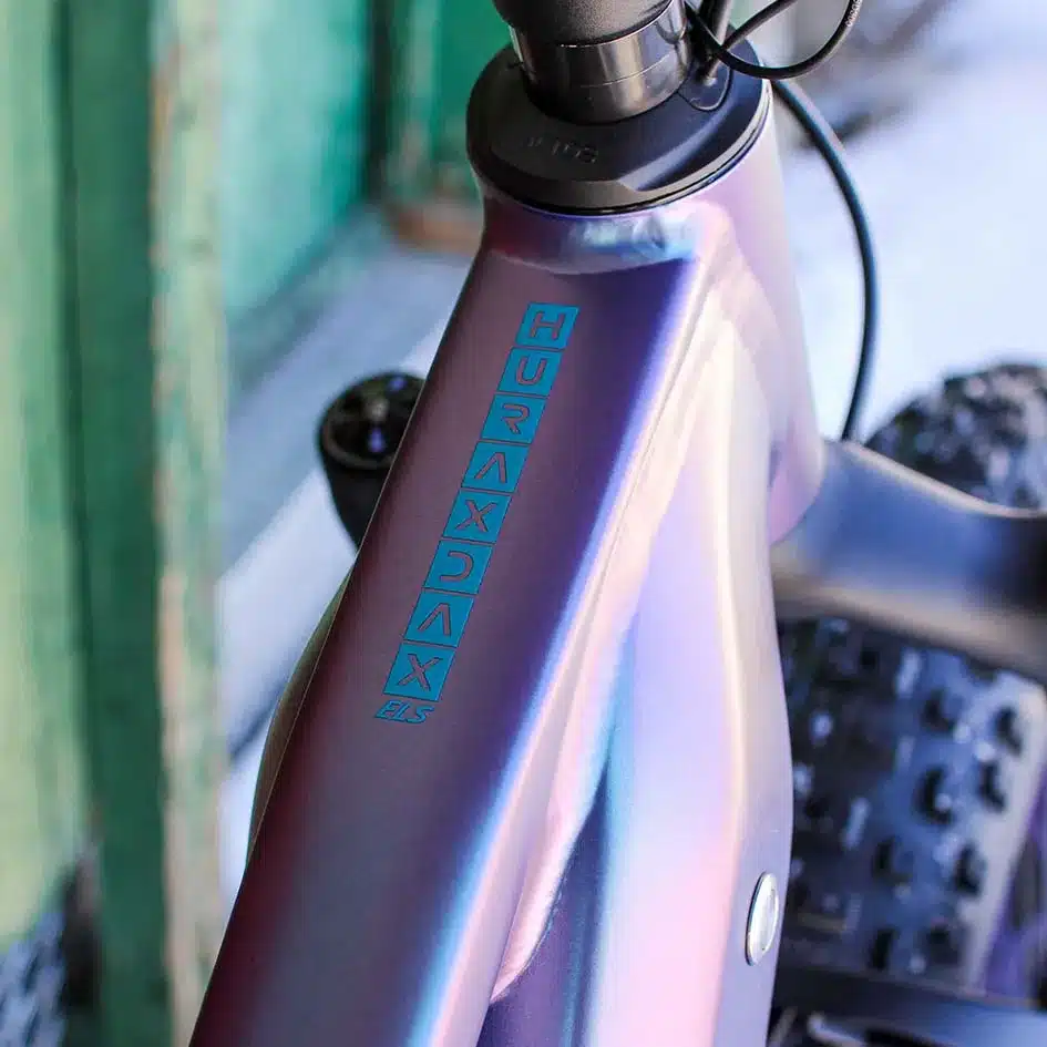 Nahaufnahme des violett-blauen Rahmens eines MAXX Huraxdax ELS X2 E-Mountainbikes mit vertikal aufgedrucktem Schriftzug des Modells.