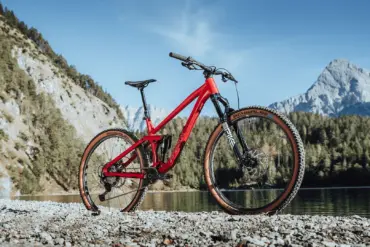 Rotes MAXX Jinxx vollgefedertes Trailbike mit 29-Zoll-Laufrädern, das auf Schotter am Ufer eines Sees mit einer beeindruckenden Berglandschaft im Hintergrund steht.