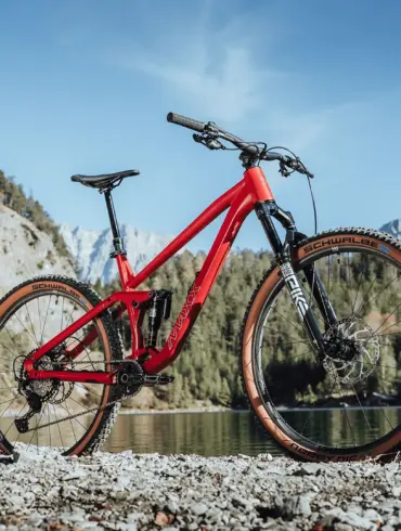 Rotes MAXX Jinxx vollgefedertes Trailbike mit 29-Zoll-Laufrädern, das auf Schotter am Ufer eines Sees mit einer beeindruckenden Berglandschaft im Hintergrund steht.