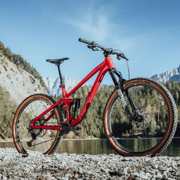 Rotes MAXX Jinxx vollgefedertes Trailbike mit 29-Zoll-Laufrädern, das auf Schotter am Ufer eines Sees mit einer beeindruckenden Berglandschaft im Hintergrund steht.