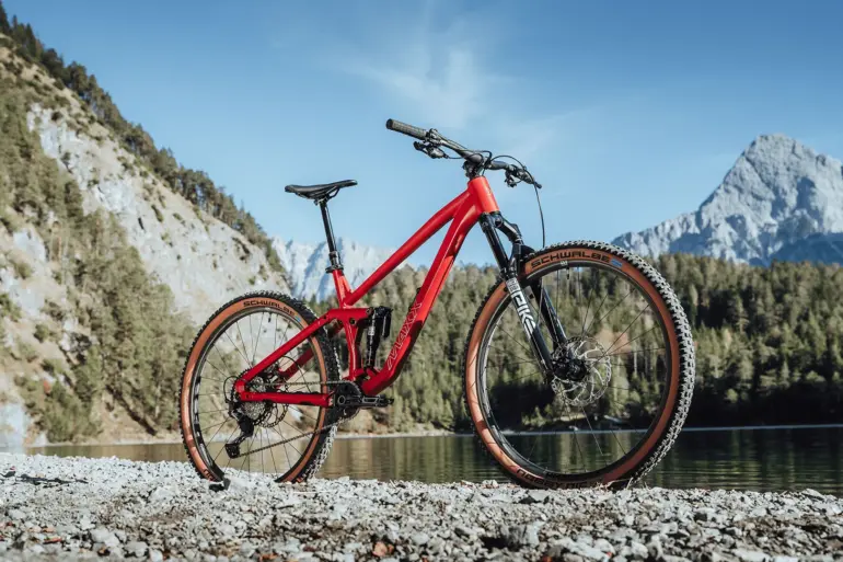 Rotes MAXX Jinxx vollgefedertes Trailbike mit 29-Zoll-Laufrädern, das auf Schotter am Ufer eines Sees mit einer beeindruckenden Berglandschaft im Hintergrund steht.