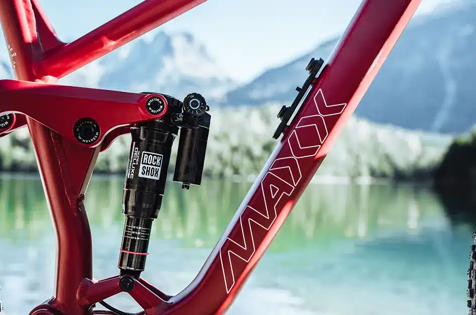 Detailaufnahme eines roten MAXX Jinxx Mountainbikes mit Fokus auf den RockShox Dämpfer und das MAXX Logo auf dem Rahmen, im Hintergrund ein See und Berge