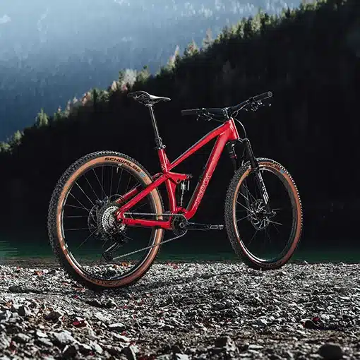Ein rotes MAXX Jinxx Mountainbike mit 29-Zoll-Laufrädern und braunen Reifen, abgestellt auf einem Kiesstrand vor einer Bergkulisse.