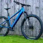 Blaues MAXX Racemaxx ELF R60 E-Mountainbike mit FOX Gabel und Schwalbe Reifen, fotografiert von der Seite vor einer Holzwand.