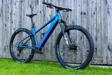 Blaues MAXX Racemaxx ELF R60 E-Mountainbike mit FOX Gabel und Schwalbe Reifen, fotografiert von der Seite vor einer Holzwand.