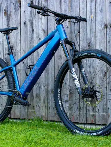 Blaues MAXX Racemaxx ELF R60 E-Mountainbike mit FOX Gabel und Schwalbe Reifen, fotografiert von der Seite vor einer Holzwand.