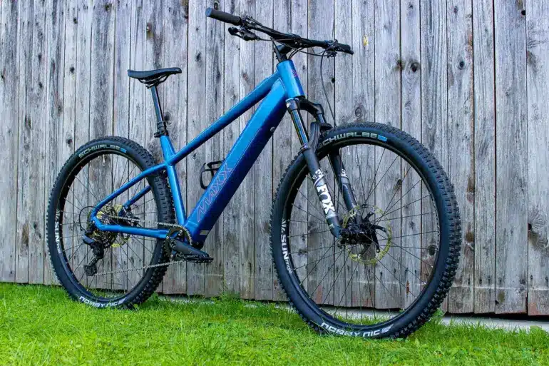 Blaues MAXX Racemaxx ELF R60 E-Mountainbike mit FOX Gabel und Schwalbe Reifen, fotografiert von der Seite vor einer Holzwand.
