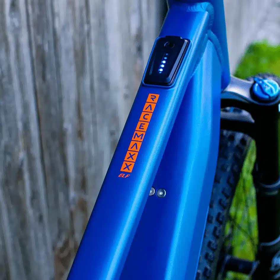 Nahaufnahme des blauen Rahmens eines MAXX Racemaxx ELF R60 E-Mountainbikes. Der Schriftzug RACEMAXX ELF ist in Orange auf dem Rahmen zu sehen. Oberhalb des Schriftzugs befindet sich die Ladeanzeige des Akkus mit leuchtenden Punkten.