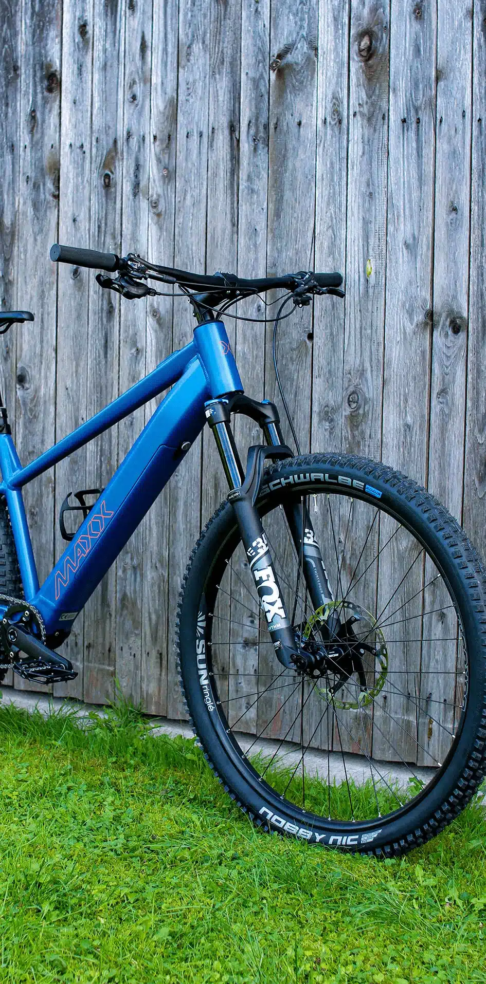 Blaues MAXX Racemaxx ELF R60 E-Mountainbike mit FOX-Federgabel und Schwalbe Reifen, aufgestellt auf grünem Gras vor einer Holzwand.