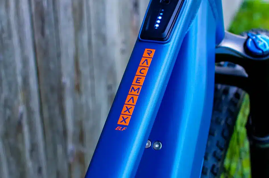 Nahaufnahme des blauen Rahmens eines E-Bikes der Marke MAXX mit dem Schriftzug RACEMAX ELF in orangefarbener Schrift, daneben eine Anzeige für den Akkustand.