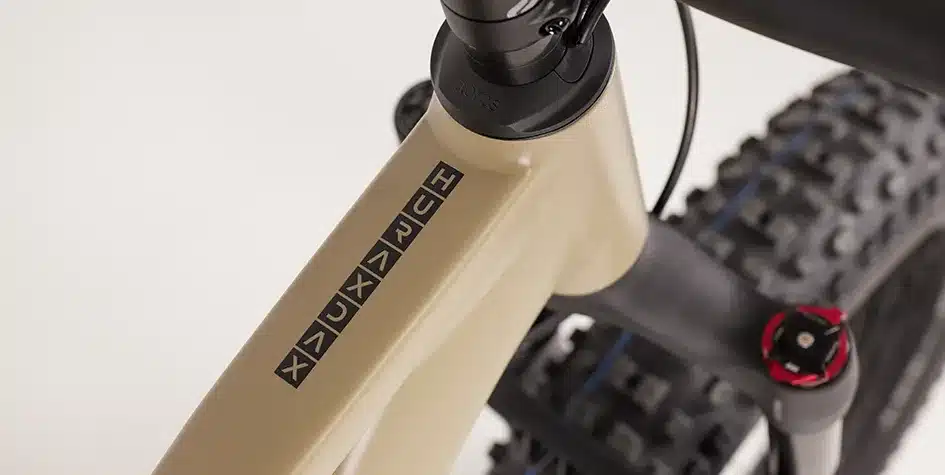 Nahaufnahme des Rahmens eines MAXX Huraxdax ELS E-Fatbikes in Beige mit dem Schriftzug 'HURAXDAX' in schwarzen Quadraten. Die Gabelkrone mit roten Akzenten und ein Teil des breiten Fatbike-Reifens sind ebenfalls sichtbar.