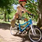 Junge fährt ein blaues woom GO 3 AUTOMAGIC Kinderfahrrad mit roten Helm auf einem gepflasterten Weg im Park unter Bäumen.