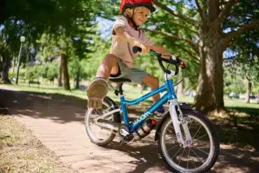 Junge fährt ein blaues woom GO 3 AUTOMAGIC Kinderfahrrad mit roten Helm auf einem gepflasterten Weg im Park unter Bäumen.