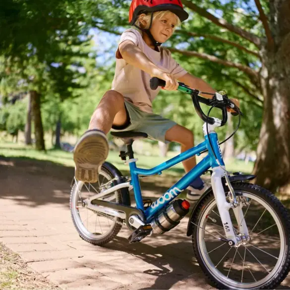 Junge fährt ein blaues woom GO 3 AUTOMAGIC Kinderfahrrad mit roten Helm auf einem gepflasterten Weg im Park unter Bäumen.