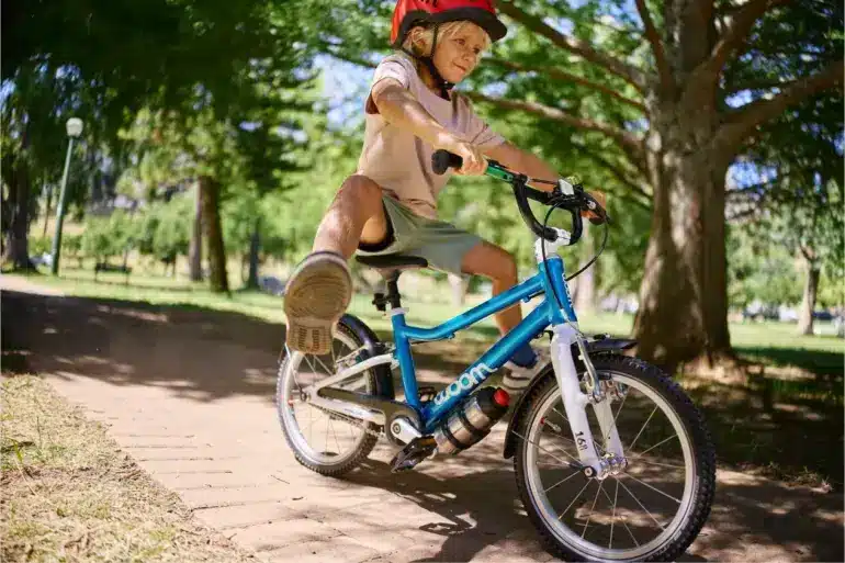 Junge fährt ein blaues woom GO 3 AUTOMAGIC Kinderfahrrad mit roten Helm auf einem gepflasterten Weg im Park unter Bäumen.