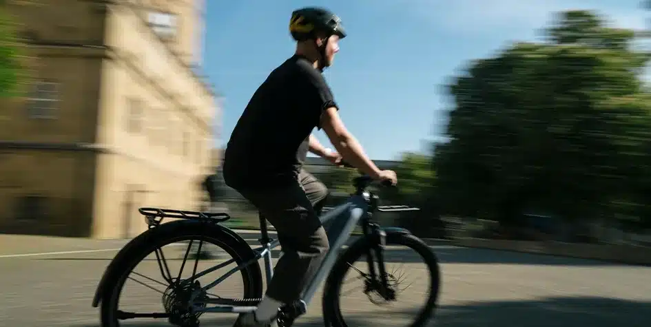Mann fährt Fahrrad mit Gepäckträger auf einer Straße in der Nähe eines Gebäudes und Bäumen.