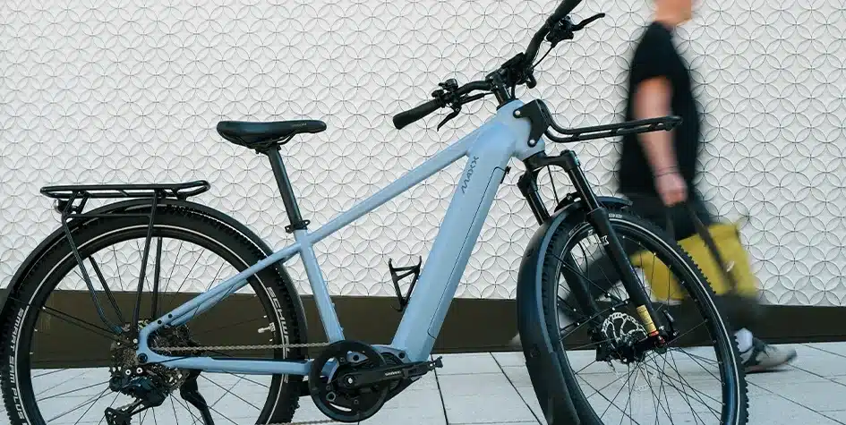 Hellblaues MAXX Pacemaxx ELS Tour E-Bike mit vollständiger Straßenausstattung wie Gepäckträger und Schutzblechen, daneben ein Mann mit gelber Tasche.
