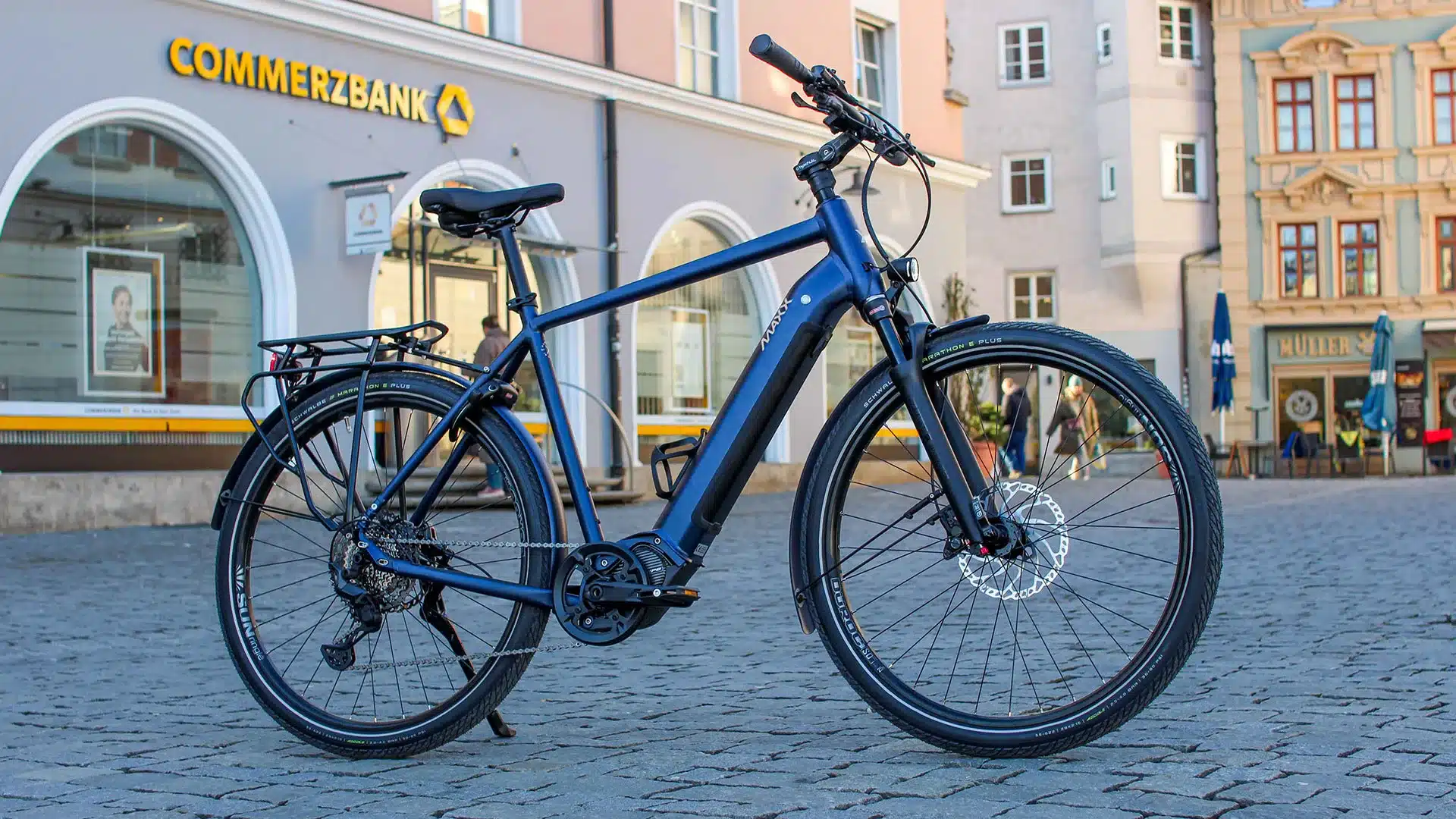 Dunkelblaues MAXX Crossmaxx ELS E-Trekkingbike mit Gepäckträger, geparkt auf einem Kopfsteinpflasterplatz vor einer Commerzbank.
