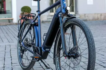 MAXX Crossmaxx ELS E-Trekkingbike, blau, mit Gepäckträger und Seitenständer, steht auf Kopfsteinpflaster.