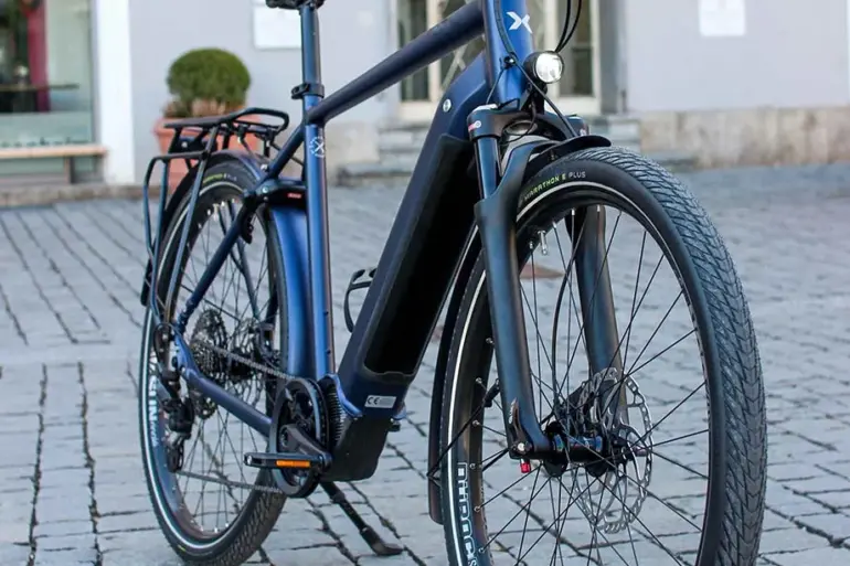 MAXX Crossmaxx ELS E-Trekkingbike, blau, mit Gepäckträger und Seitenständer, steht auf Kopfsteinpflaster.