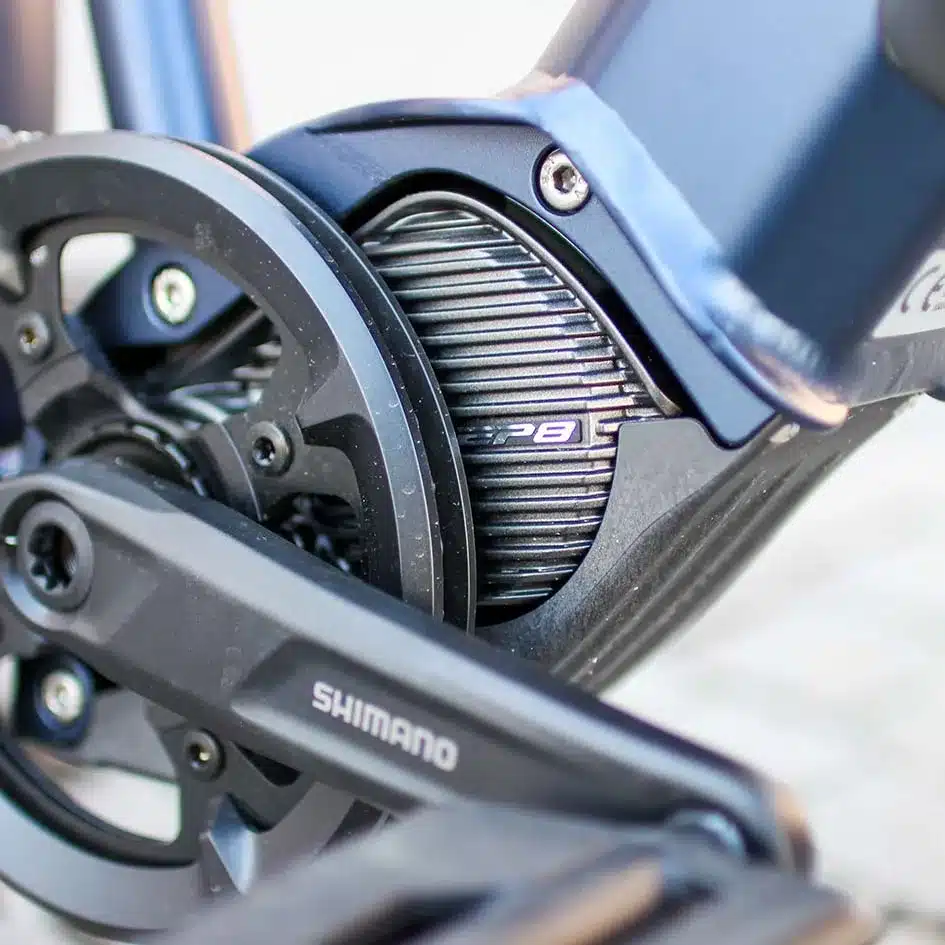 Nahaufnahme des schwarzen Shimano EP8 Mittelmotors, integriert in den dunkelblauen Rahmen eines MAXX Crossmaxx ELS E-Trekkingbikes, mit schwarzem Kurbelarm und Kettenblatt von Shimano.