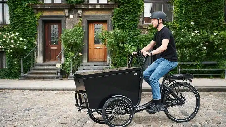 Ein Mann fährt ein schwarzes Winther CX Dreirad-Lastenrad auf einer Kopfsteinpflasterstraße.