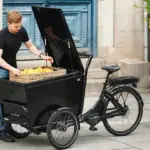 Ein Mann mit schwarzem T-Shirt und Jeans belädt ein dreirädriges, schwarzes Winther CX Lastenrad mit einer Holzkiste, die mit gelben Zitronen gefüllt ist.