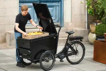Ein Mann mit schwarzem T-Shirt und Jeans belädt ein dreirädriges, schwarzes Winther CX Lastenrad mit einer Holzkiste, die mit gelben Zitronen gefüllt ist.