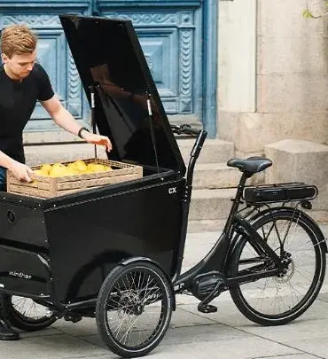 Ein Mann mit schwarzem T-Shirt und Jeans belädt ein dreirädriges, schwarzes Winther CX Lastenrad mit einer Holzkiste, die mit gelben Zitronen gefüllt ist.