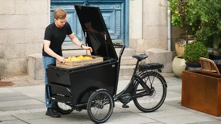 Ein Mann mit schwarzem T-Shirt und Jeans belädt ein dreirädriges, schwarzes Winther CX Lastenrad mit einer Holzkiste, die mit gelben Zitronen gefüllt ist.
