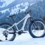 Ein weißes E-Fatbike MAXX Jagamoasta ELS SH steht in tiefer Schneedecke. Im Hintergrund ist eine verschneite Bergkulisse mit Nadelbäumen zu sehen.