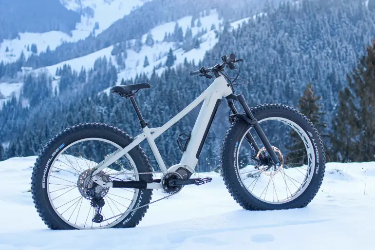 Ein weißes E-Fatbike MAXX Jagamoasta ELS SH steht in tiefer Schneedecke. Im Hintergrund ist eine verschneite Bergkulisse mit Nadelbäumen zu sehen.