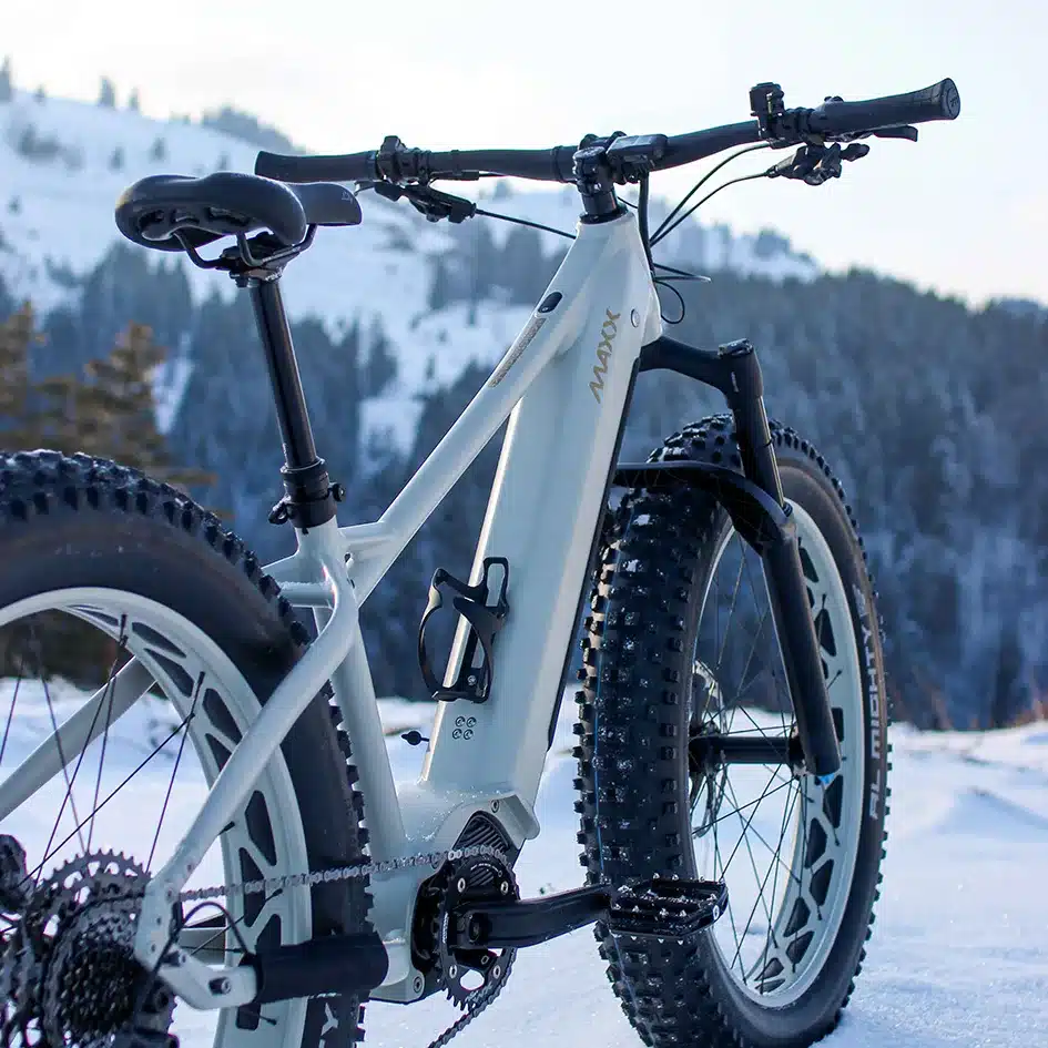 Detailansicht eines MAXX Jagamoasta ELS SH E-Fatbikes mit breiten Reifen und weißem Rahmen, geparkt in einer winterlichen Schneelandschaft mit verschneiten Bergen im Hintergrund.