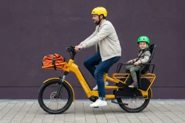 Ein Mann fährt ein gelbes Maxx Maxximo Urban Longtail-E-Bike mit einem Kind, das in einem Kindersitz auf dem Gepäckträger sitzt. Der Mann trägt einen gelben Helm, das Kind trägt einen grünen Helm. Vorne am Rad ist eine orangefarbene Tasche befestigt.