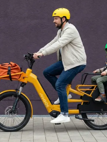 Ein Mann fährt ein gelbes Maxx Maxximo Urban Longtail-E-Bike mit einem Kind, das in einem Kindersitz auf dem Gepäckträger sitzt. Der Mann trägt einen gelben Helm, das Kind trägt einen grünen Helm. Vorne am Rad ist eine orangefarbene Tasche befestigt.