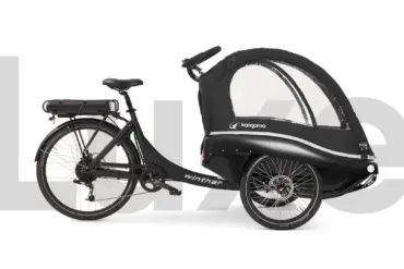 Kangaroo Luxe Lastenrad, ein schwarzes Elektro-Dreirad mit geschlossener Transportkabine für Kinder und Gepäck, mit Gepäckträger und Batterie.