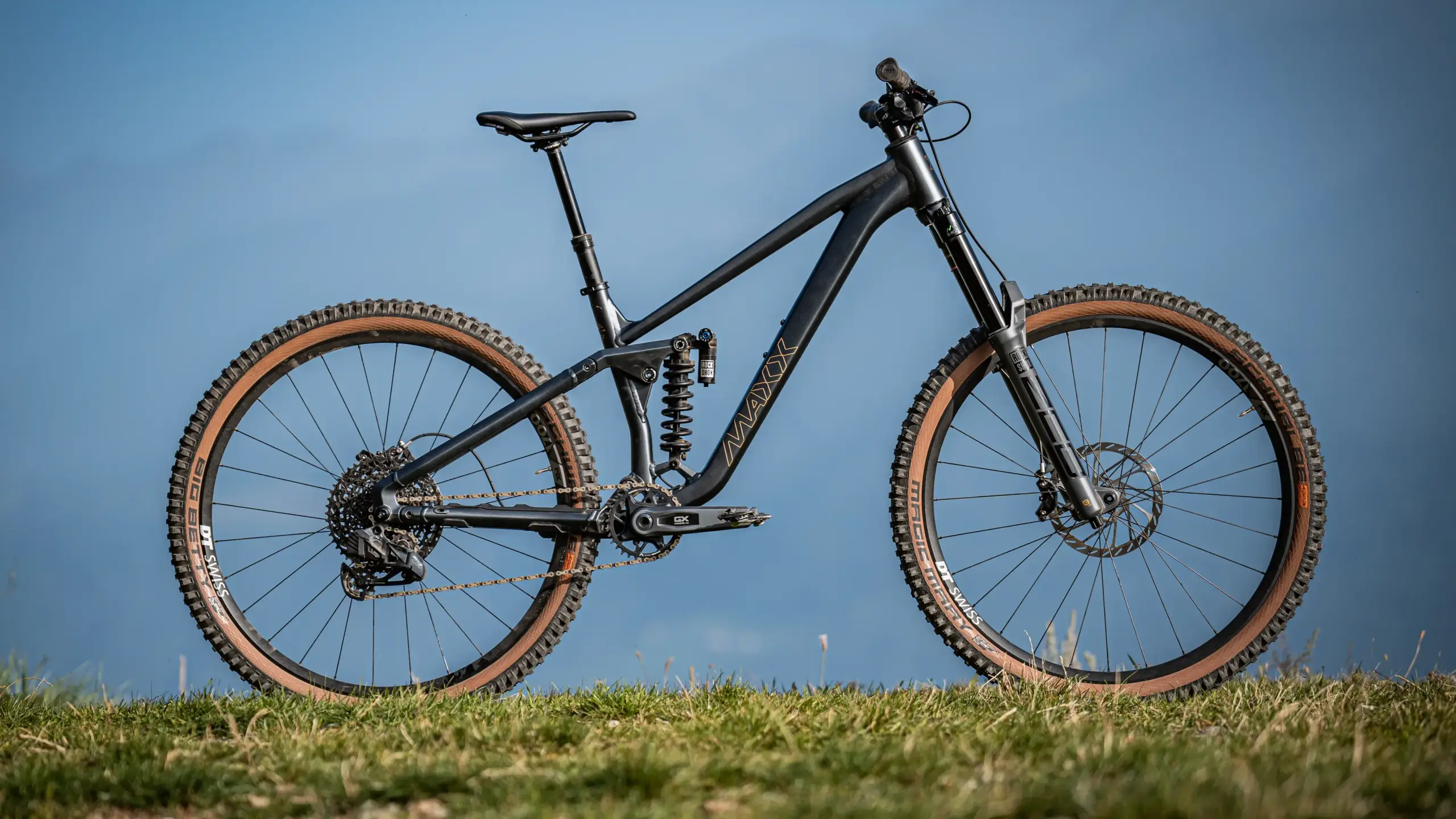 Ein schwarzes MAXX Fab4 29 Zoll Enduro Mountainbike mit braunen Reifen, voller Federung und Shimano Komponenten, abgebildet auf Gras.
