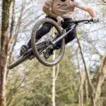 Ein Mann mit Helm und Sonnenbrille fährt mit einem Mountainbike durch die Luft auf einem Waldtrail.