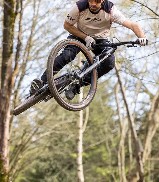 Ein Mann mit Helm und Sonnenbrille fährt mit einem Mountainbike durch die Luft auf einem Waldtrail.