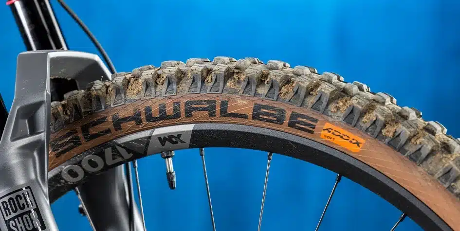 Detailansicht eines Schwalbe Mountainbike-Reifens mit brauner Karkasse und groben Stollen, montiert auf einer Felge. Die RockShox Federgabel ist angedeutet.