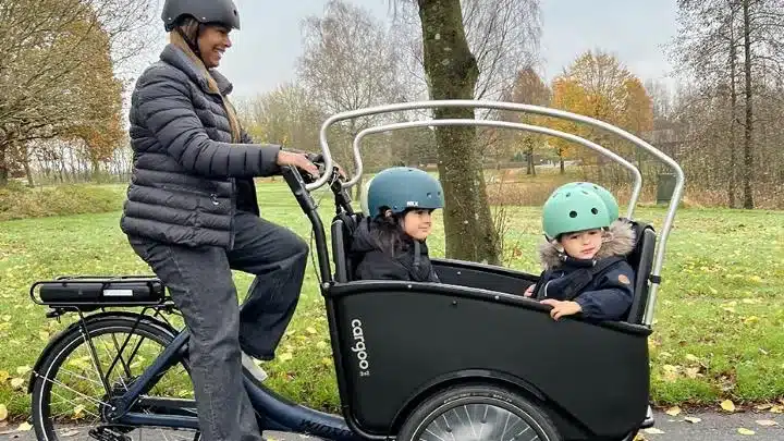 Frau fährt ein schwarzes Winther Cargoo 2+2 Lastenrad mit zwei Kindern (Junge und Mädchen) in Helmen auf dem Sitzplatz. Das Rad befindet sich auf einem Feldweg in einem Park mit Bäumen und Laub.