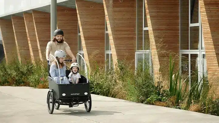 Mann fährt ein dreirädriges Cargoo Lastenrad mit einer Frau auf dem Beifahrersitz und einem Kind in der Kabine.