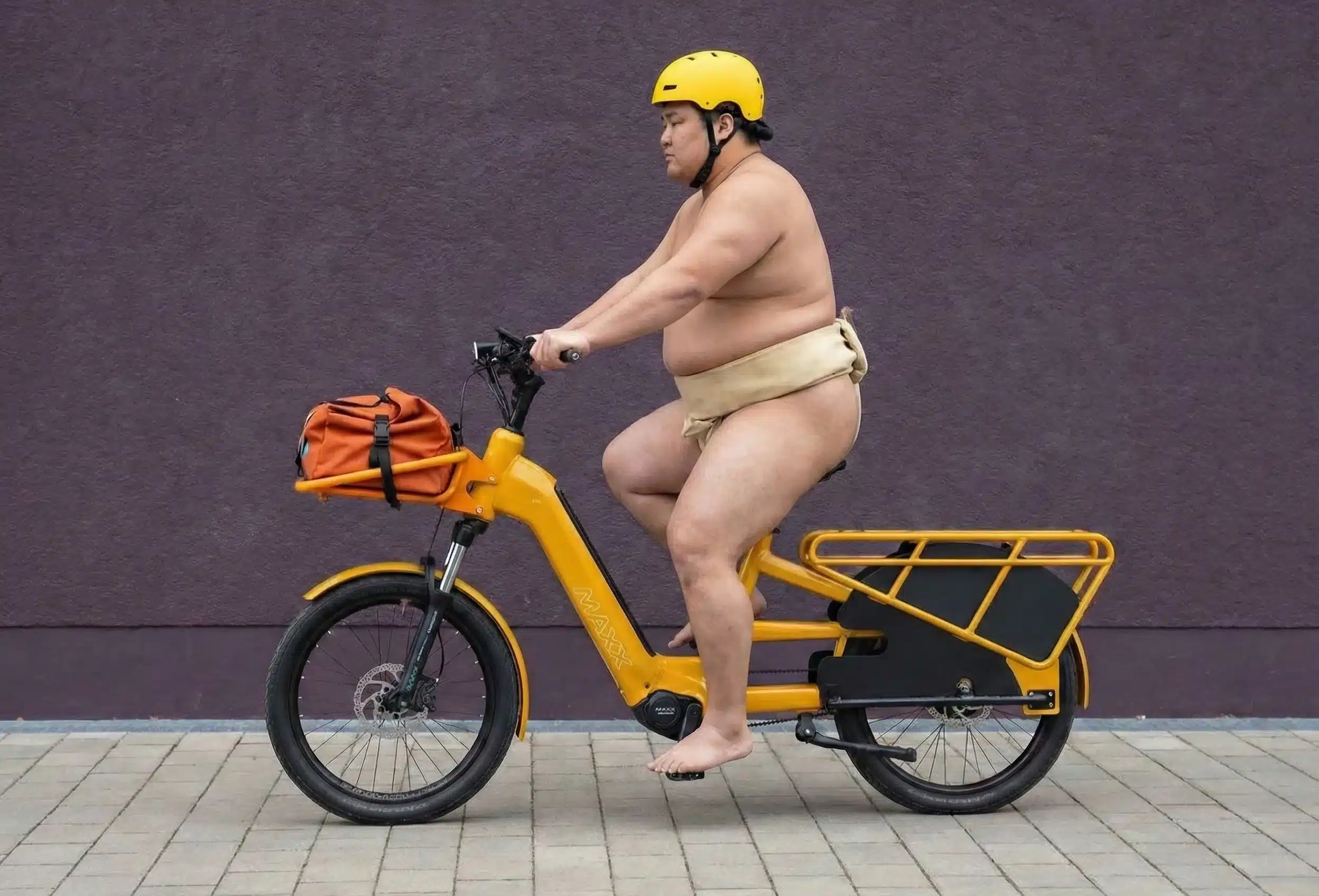Ein asiatisch aussehender Mann mit gelbem Helm und einem traditionellen Sumo-Lendenschurz fährt ein gelbes E-Bike des Modells MAXX Maxximo Strong. Das Fahrrad hat einen orangefarbenen Gepäckbeutel auf dem vorderen Gepäckträger. Der Hintergrund ist eine dunkle, texturierte Wand.