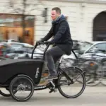 Mann fährt ein schwarzes Winther Cargoo Box Dreirad-Lastenrad mit überdachter Ladebox auf einer belebten Straße, umgeben von geparkten Fahrrädern und Autos im Hintergrund.