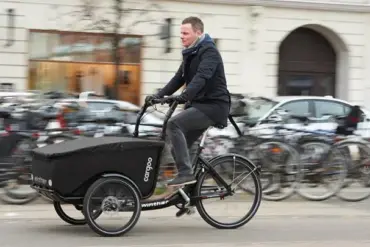 Mann fährt ein schwarzes Winther Cargoo Box Dreirad-Lastenrad mit überdachter Ladebox auf einer belebten Straße, umgeben von geparkten Fahrrädern und Autos im Hintergrund.
