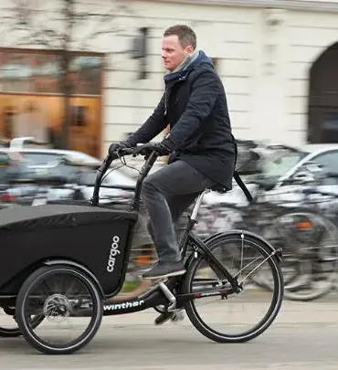 Mann fährt ein schwarzes Winther Cargoo Box Dreirad-Lastenrad mit überdachter Ladebox auf einer belebten Straße, umgeben von geparkten Fahrrädern und Autos im Hintergrund.