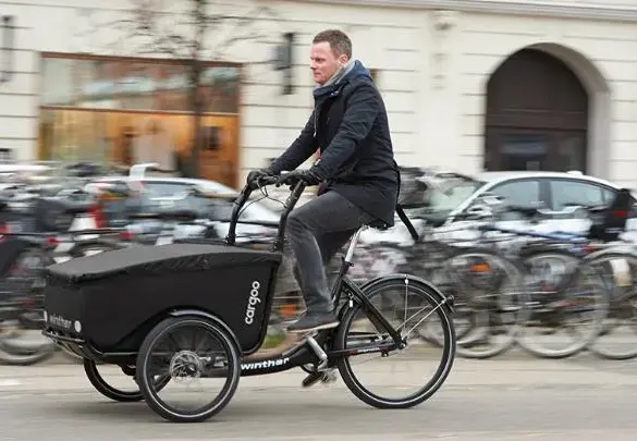 Mann fährt ein schwarzes Winther Cargoo Box Dreirad-Lastenrad mit überdachter Ladebox auf einer belebten Straße, umgeben von geparkten Fahrrädern und Autos im Hintergrund.