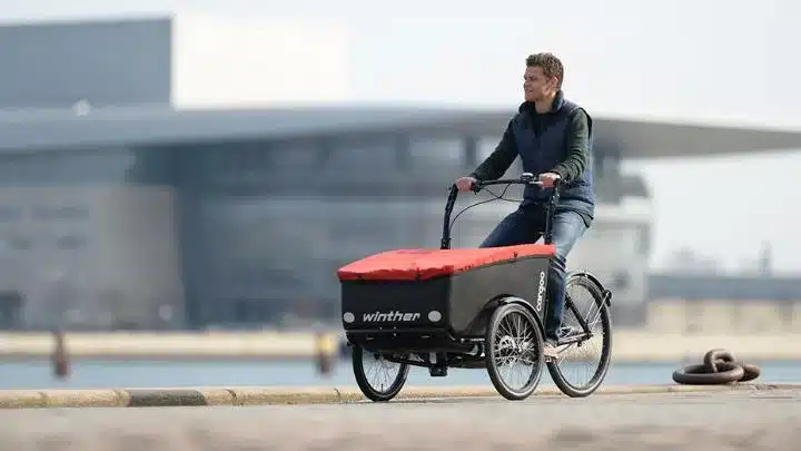 Ein Mann fährt ein schwarzes Winther Cargoo Box Dreirad-Lastenrad mit roter Abdeckung über die Ladebox.