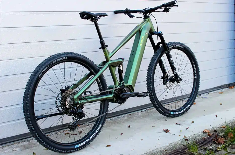 Ein Maxx Tourmaxx ELS Sport E-Mountainbike in Grün steht auf einem Betonboden vor einer weißen Garagentür. Das Fahrrad ist voll gefedert, hat 29-Zoll-Laufräder und eine Shimano-Schaltung.